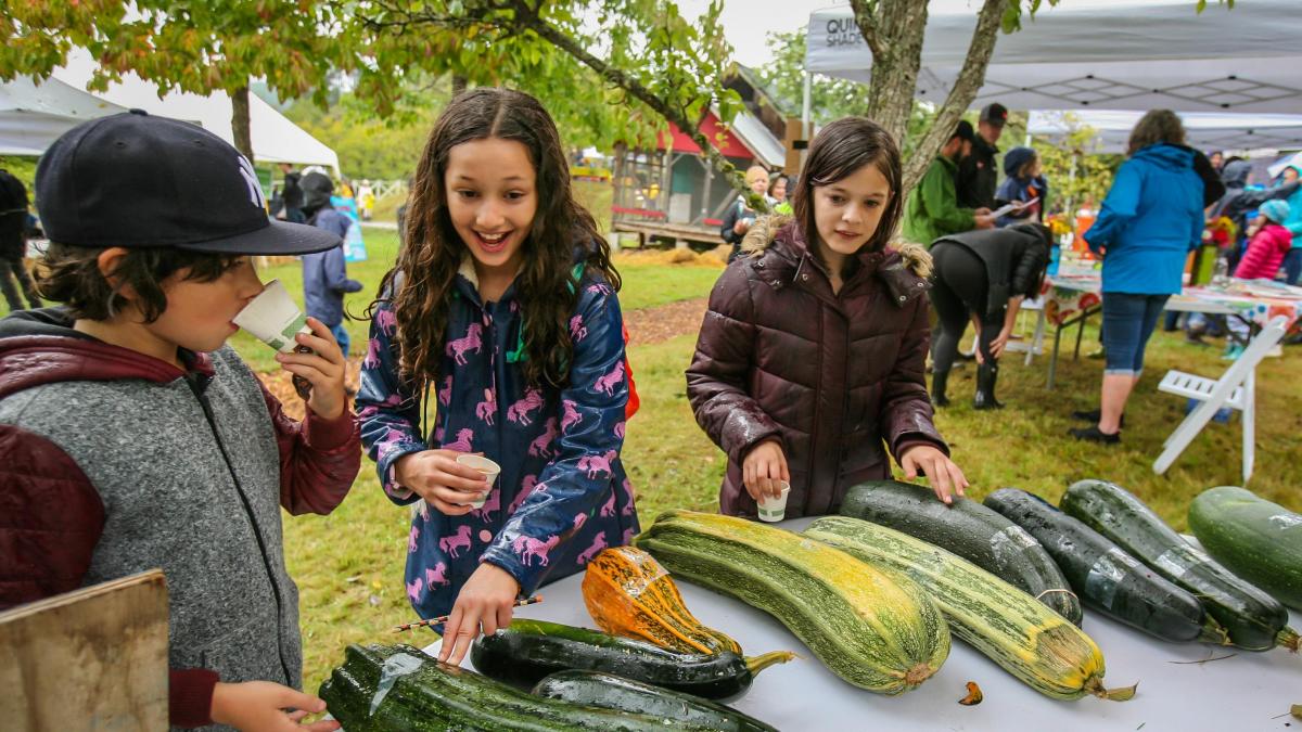 Three children examining a selection of large gourds at an outdoor fair, with one child visibly excited and pointing, while the others taste or inspect the produce.